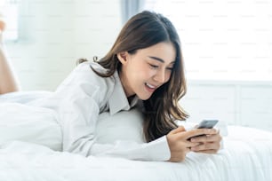Asian attractive woman use mobile phone chat on bed at home in morning. Happy casual beautiful female lying down in bedroom enjoy holiday weekend, use smartphone communicate with technology in house.