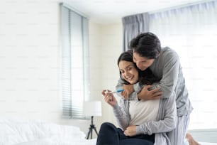 Asian young couple feeling happy after look at positive pregnancy test. Beautiful loving woman surprises boyfriend with good news to be future parents on bed in bedroom. Family-Expecting baby concept.