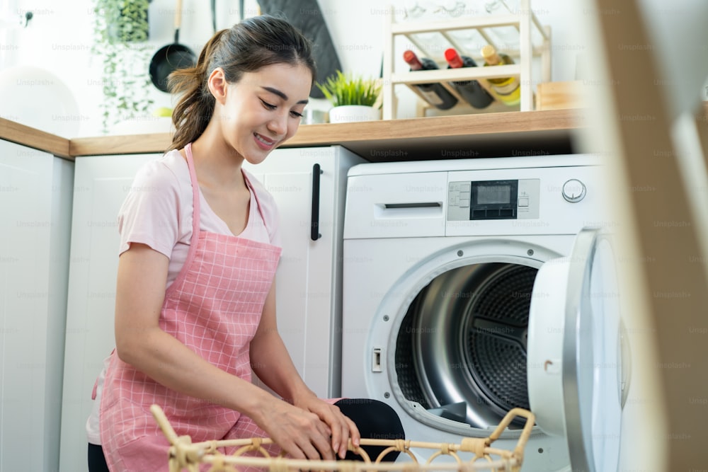 Asian beautiful woman put dirty clothes to washing machine in house. Attractive girl wear apron sit on floor, feel happy to loads laundry in washer appliance at home. Domestic-House keeping concept.