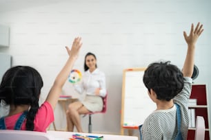 Caucasian beautiful young woman teacher teaching art to kids at school. Rear view of group young little kid student raise hand to answer questions with confident in schoolroom enjoy to study at school