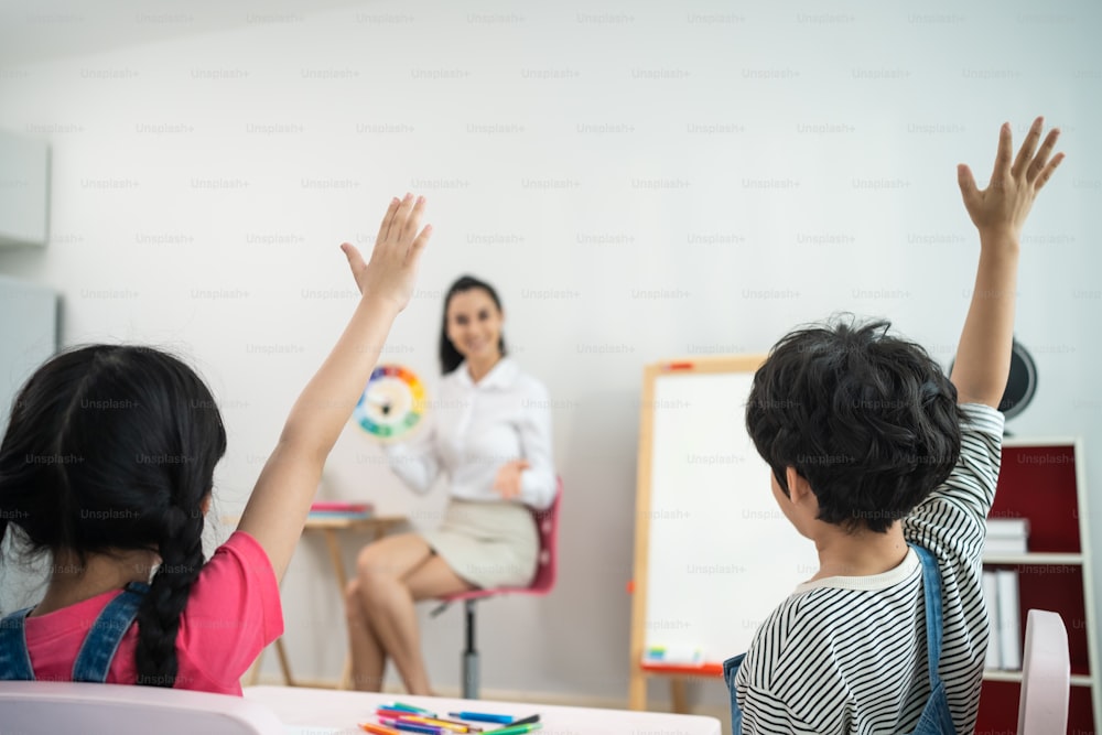 Caucasian beautiful young woman teacher teaching art to kids at school. Rear view of group young little kid student raise hand to answer questions with confident in schoolroom enjoy to study at school