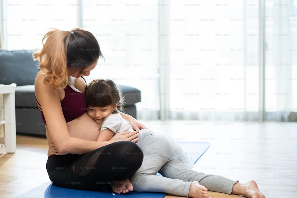 Asian young lovely daughter hugging beautiful pregnant mother tummy. Attractive expecting pregnancy girl embrace little girl with happiness and love while doing yoga exercise in living room at home.