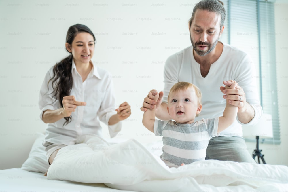Caucasian loving parents play with baby boy child on bed in bedroom. Happy family, attractive beautiful young couple dad and mom dance with cute infant toddler son enjoy morning activity after wake up