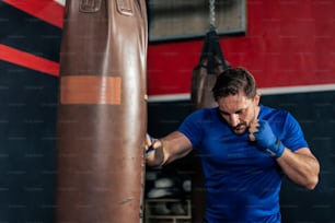 Portrait Caucasian professional sport young man exercising in fitness. Attractive athlete male in sportswear and boxing gloves workout by punching sand bag to maintain strong muscle in fitness club.