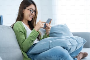 Asian excited young woman winner feeling happy while looking at phone. Attractive girl student sit on sofa in living room and found out good news on smartphone then celebrate online success in house.