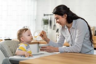 Caucasian attractive parents feeding foods to baby toddler in kitchen. Happy family, loving mother cook and serve healthy foods to little cute kid  boy son infant while sit on chair for lunch in house
