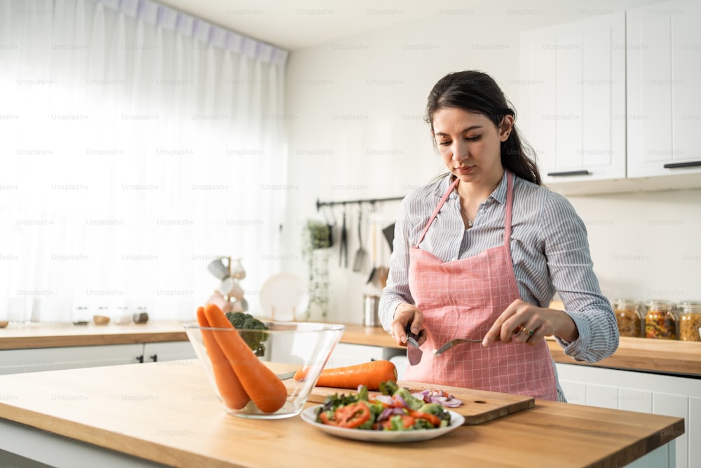Caucasian loving woman wear apron cook green salad in kitchen at home. Young beautiful girl feeling happy and enjoy eating vegetables healthy foods to diet and lose weight for health care in house.