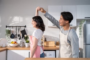Asian attractive loving couple enjoy dance together in kitchen at home. Beautiful romantic new marriage man and woman wear aporn feeling happy and enjoy activity relationship in house listen to music.