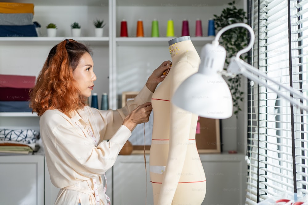 Asian beautiful tailor woman working on clothes in tailoring atelier. Attractive young girl fashion designer use tape measure new handmade suit on mannequin in workshop room. Fashion business concept.