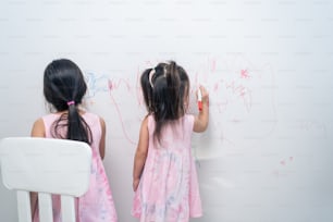Asian young sibling kid girl enjoy paint on white wall in living room. Little adorable children having fun drawing and coloring art picture with happiness enjoy creativity activity on holiday at home.
