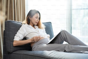 Asian young beautiful pregnant woman applying headphones on her belly. Attractive expectant pregnancy girl mother sit on sofa indoors in living room and play music for newborn baby in belly to listen.