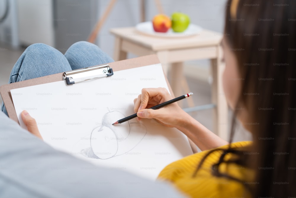 Close up of talented woman artist drawing picture on painting board. Attractive beautiful girl lying on sofa, feel happy to creating artwork by sketching out an apple enjoy creativity activity at home