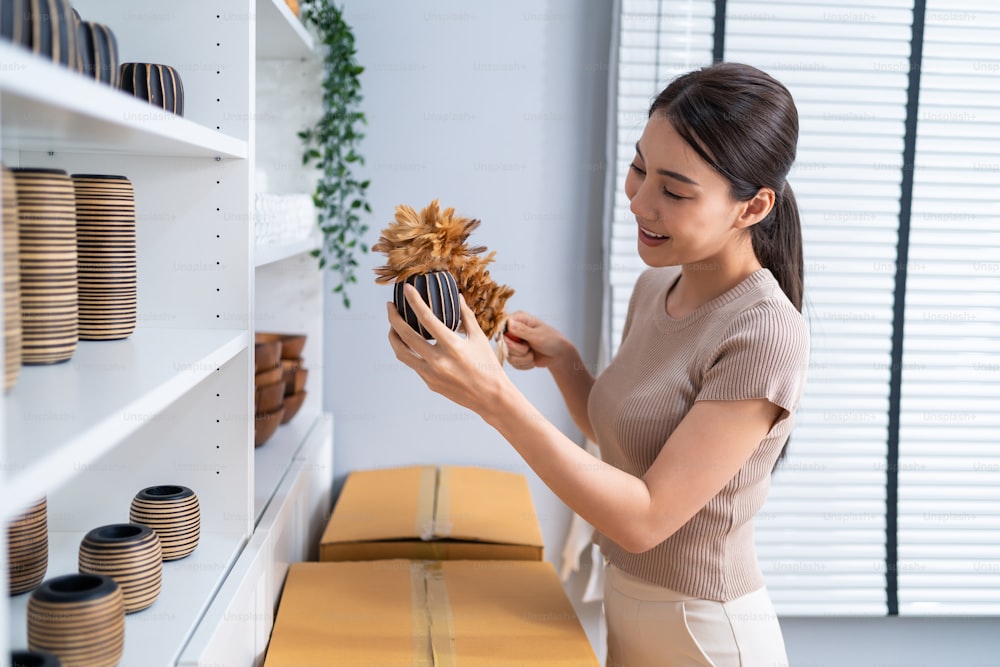 Asian beautiful woman clean vase goods order for customer from shelf. Young attractive business girl working to preparing parcel boxes checking ecommerce shipping online retail to sell at home store.