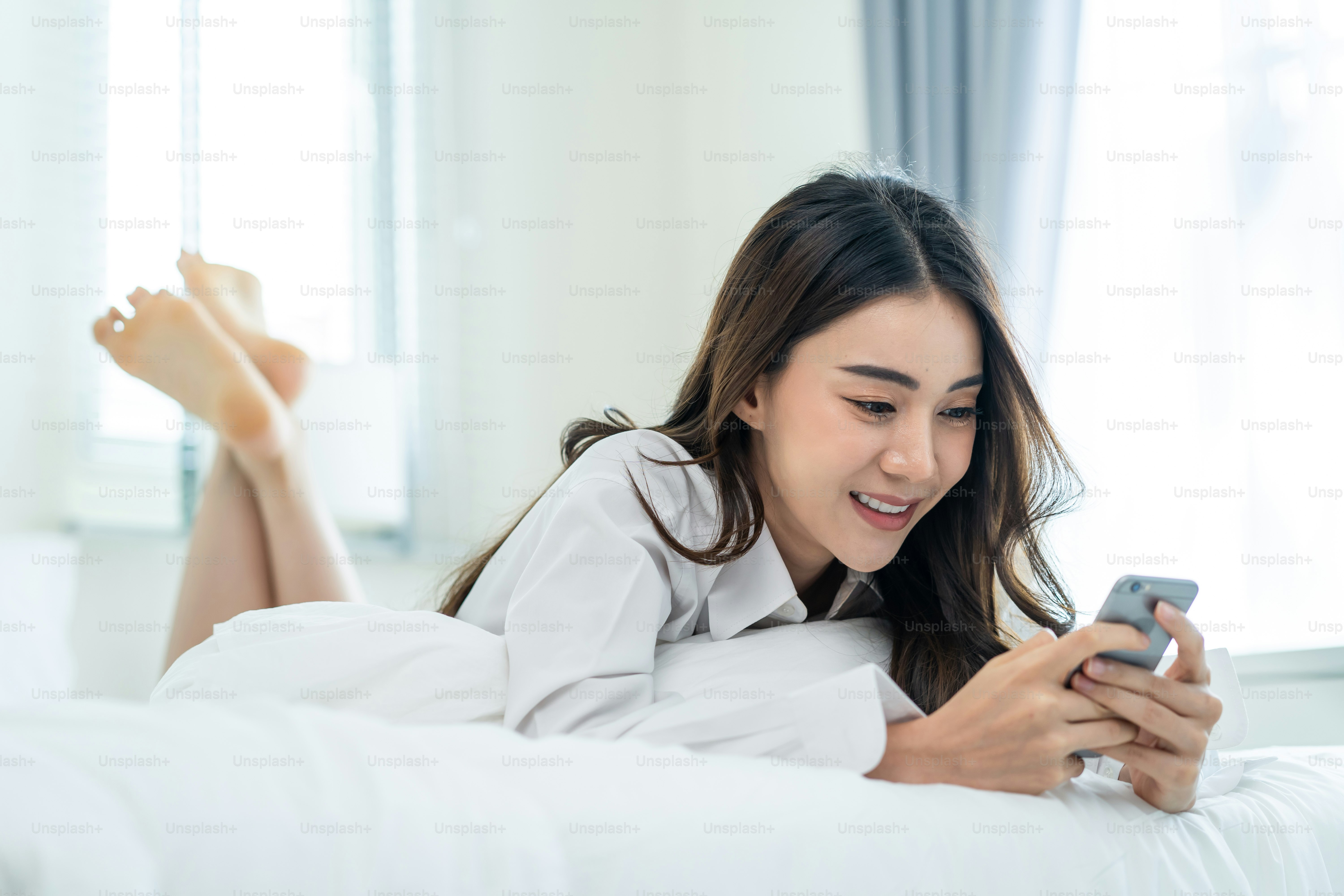 Mulher atraente asiática usar bate-papo de telefone celular na cama em casa de manhã. Feliz casual bela fêmea deitada no quarto desfrutar de fim de semana de férias, usar smartphone se comunicar com a tecnologia em casa.