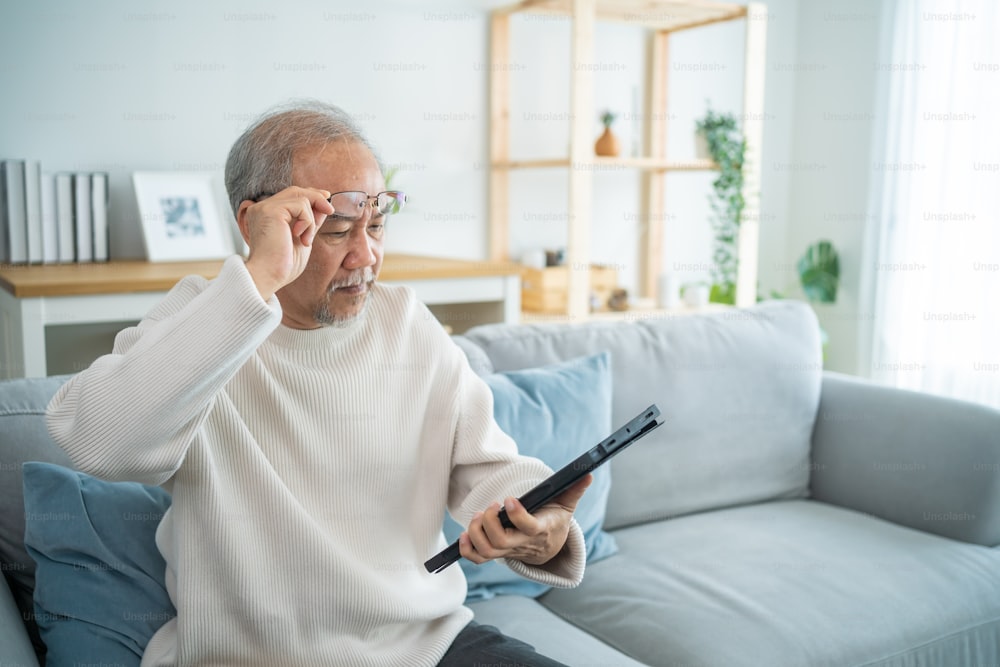 Asian senior elderly male using digital tablet in living room at home. Strong older mature grandfather wear eyeglasses, sitting on sofa and chat on laptop communicate with family enjoy retirement life