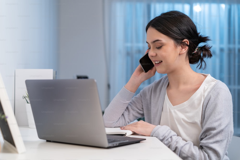 Asian young beautiful businesswoman working at home during night time. Attractive freelancer girl sit on study table, use mobile phone call for late work meeting and write note in living room in house