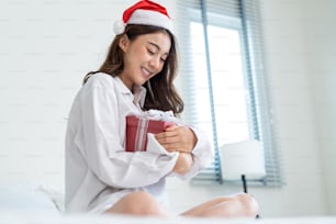 Asian beautiful young woman wear Santa hat, open xmas present gift box. Attractive girl feeling happy and excited for surprise from family and ready to celebrate holiday Christmas together in house.