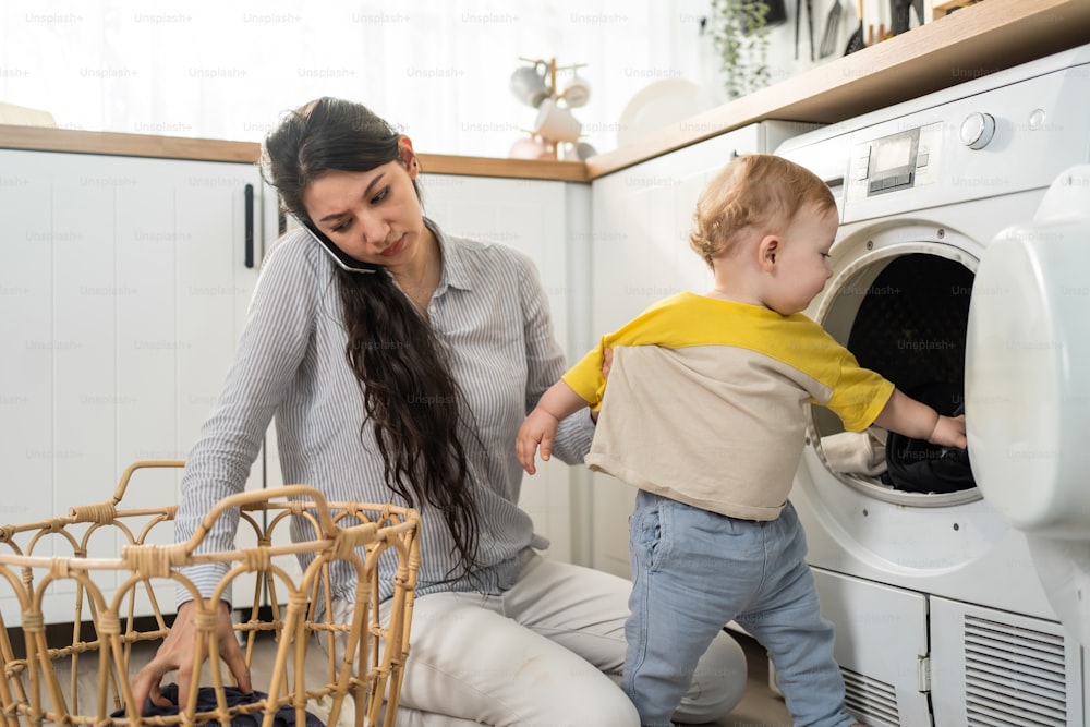 Madre caucásica ocupada haciendo tareas domésticas con un bebé pequeño en la cocina. La hermosa mamá usa la llamada telefónica para el trabajo y pone la ropa en la lavadora con su hijo jugando en la casa. Tareas domésticas familiares.