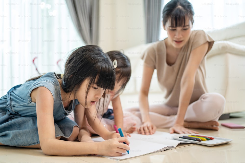 Asian Little siblings girl draw and color picture with mother on floor. Beautiful loving parent, mom spend time with small cute kid daughters enjoy activity at house. Parenting relationship concept.
