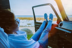 アジアの女性は自然を旅します。夏のビーチでのんびりと旅を。車に座って海を見て幸せで、車の外に足があります。