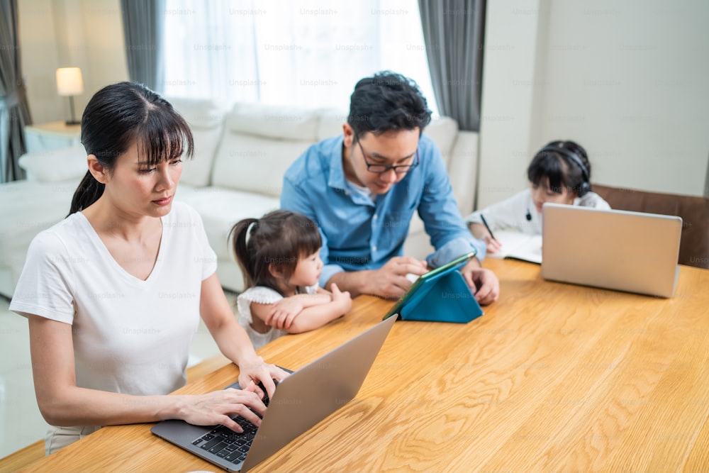 Asian busy family stay home Parents work at home with daughters beside. Loving Father and mother help and support little kid girl doing remote online study by remote meeting during lockdown in house.