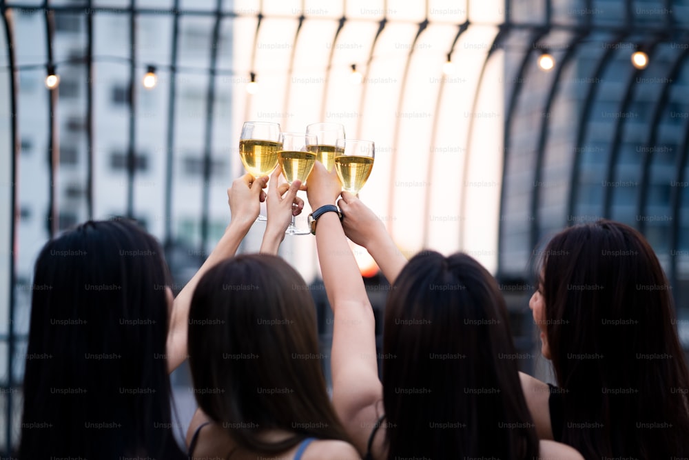 Group of young Asian cute women having party at rooftop restaurant. Girls drinking white wine alcohol and toasting glass feeling happy enjoy, fun seen from behind. Night lifestyle concept.