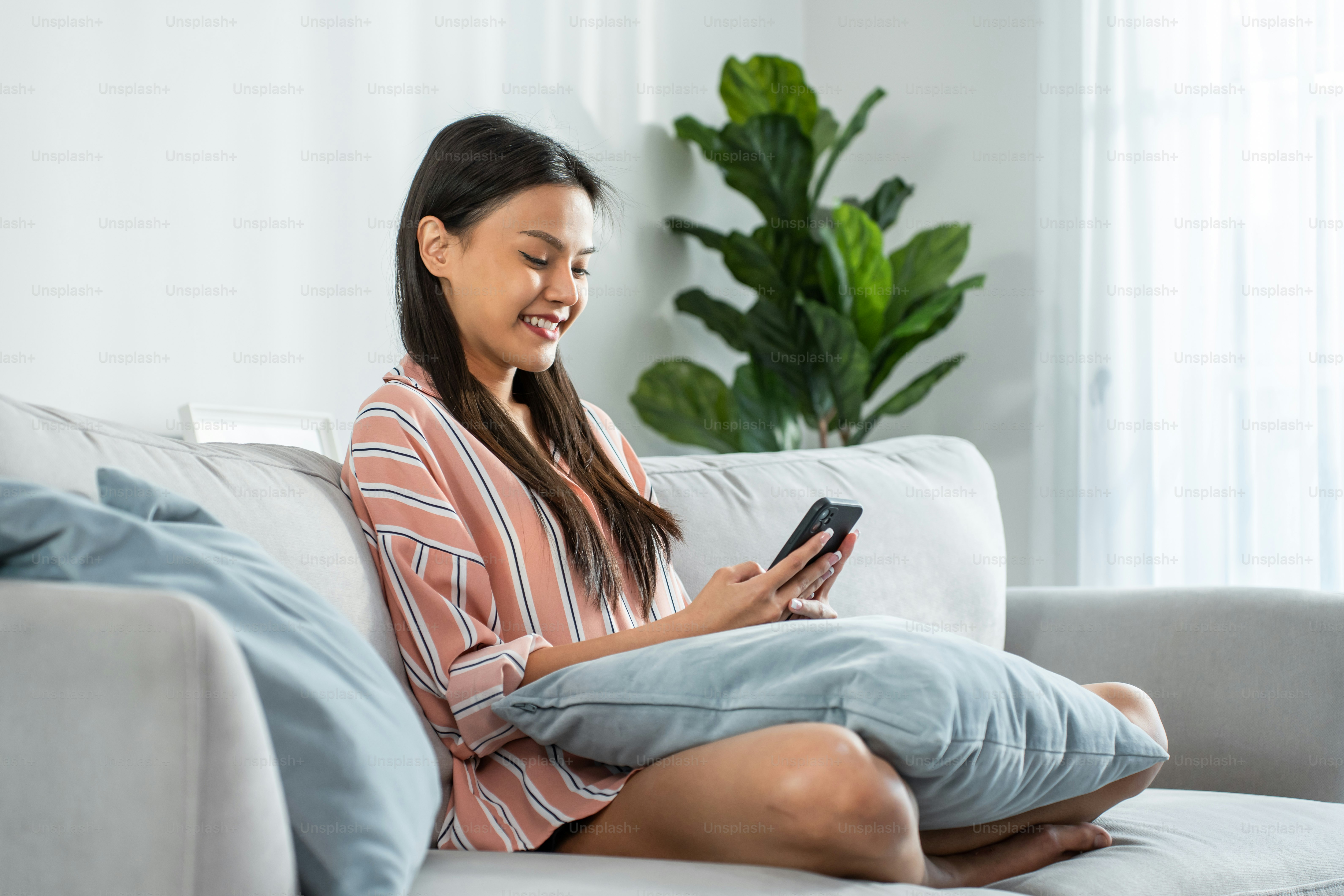 Belle femme asiatique s’assoit sur le canapé et discute sur le téléphone portable dans la maison. Heureuse jeune fille attrayante passer du temps libre à la maison, se sentir détendue et profiter de communiquer et discuter sur smartphone dans le salon.