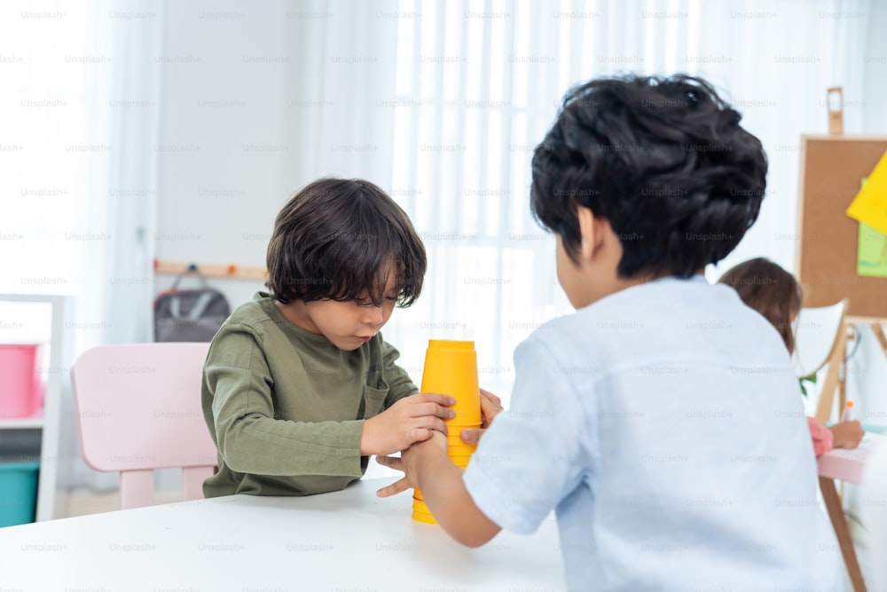Asian two little friends kid boy playing toy together in schoolroom. Two child student enjoy, spending time play toy stacks cup while during break with feeling fun together in classroom at school.