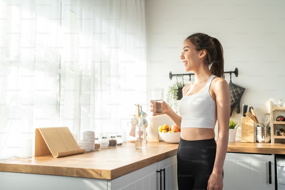 Asian beautiful woman in sportswear drink water after exercise at home. Young thirsty active sport girl takes a sips of clean mineral natural in cup after workout for health care in kitchen in house.