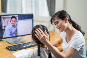 Homeschool Asian little young girl learning online class from school teacher by digital remote internet meeting due to coronavirus pandemic. Kid looking computer and writing note, sitting with mother.