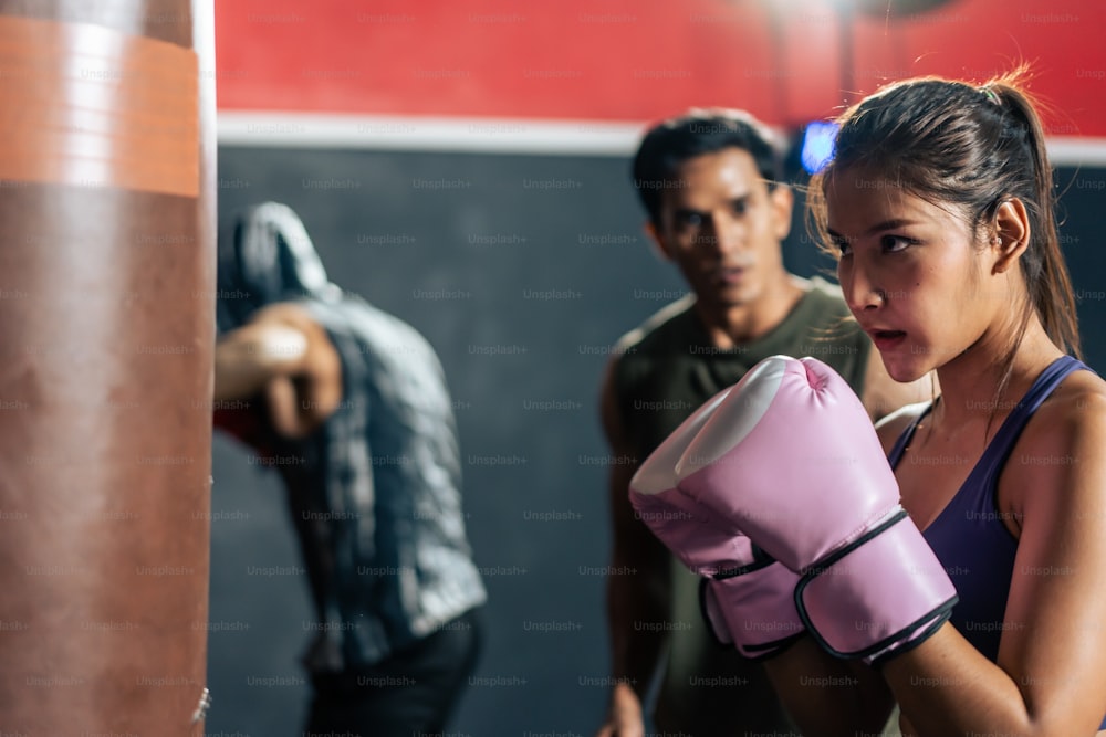 Group of athlete hitting a punching bag or sandbag at fitness gym club. Asian Fitness trainer instructor and young beautiful active sportsman and woman exercising doing boxing or Muay Thai in stadium.