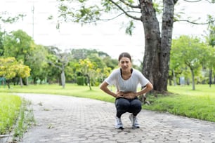 アジアの魅力的なスポーツの女性は、通りを走った後、胃の痛みを感じます。スポーツウェアを着た美しい女の子は、公共の公園で夕方に屋外でジョギングトレーニングをして運動しながら腹痛の病気を患っています