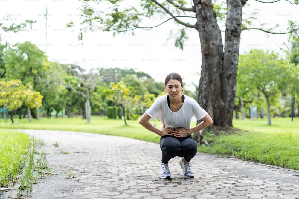 Asian attractive sport woman feels pain on stomach after run on street. Beautiful girl in sportswear having stomach ache illness while exercise by jogging workout outdoor in the evening in public park
