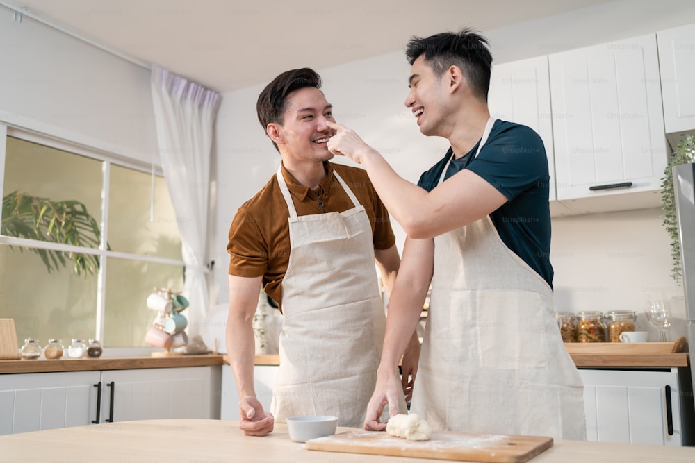 Giovane famiglia gay maschio LGBTQ asiatica si diverte a cuocere la panetteria in cucina a casa. Attraente bella coppia romantica dell'uomo indossa il grembiule sentendosi felice e allegro per trascorrere il tempo cucinando i cibi insieme in casa.