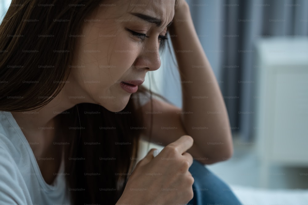 Asian beautiful depression girl crying in tears sit on bed in bedroom. Attractive unhappy young woman feeling sad lonely and upset with life problem and hold tissue on hands in dark night room at home