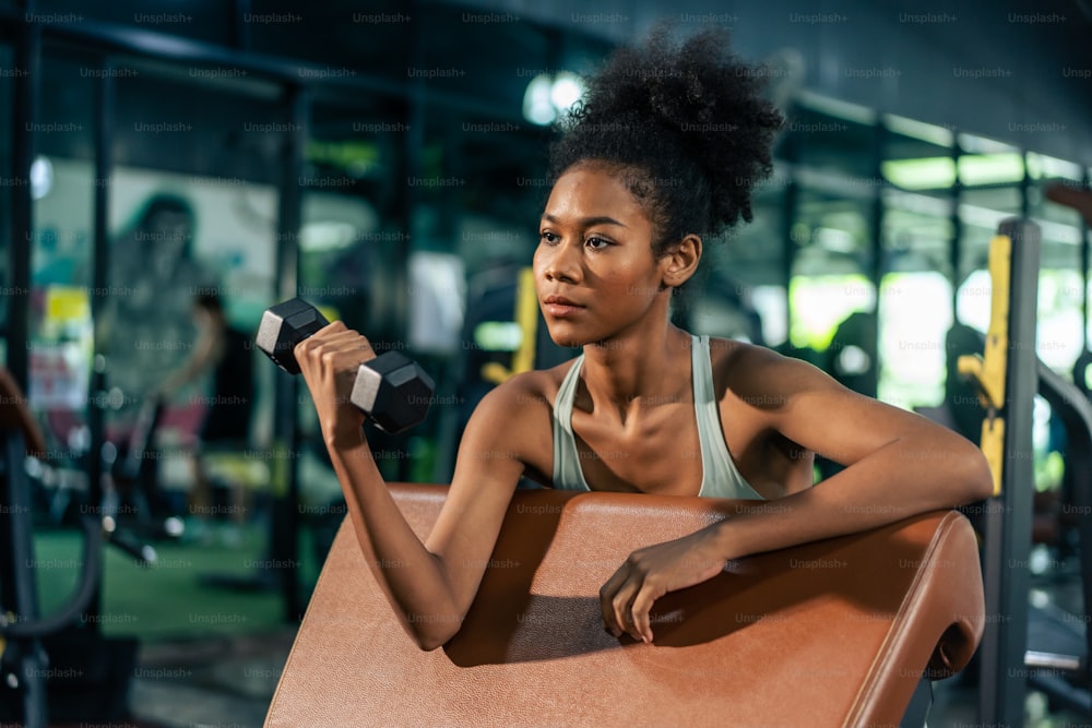 Young beautiful African american sportswoman exercise in gym stadium. Attractive active athlete girl in sportswear workout by lifting weight dumbbells to build muscle for health care and at fitness.