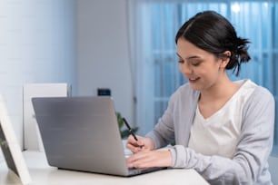 Une jeune femme asiatique étudie virtuellement en ligne et travaille dans le salon à la maison. Apprentissage à distance de belle fille d’affaires tapant sur un ordinateur portable la nuit dans la maison. L’école à la maison en retard du concept de travail.