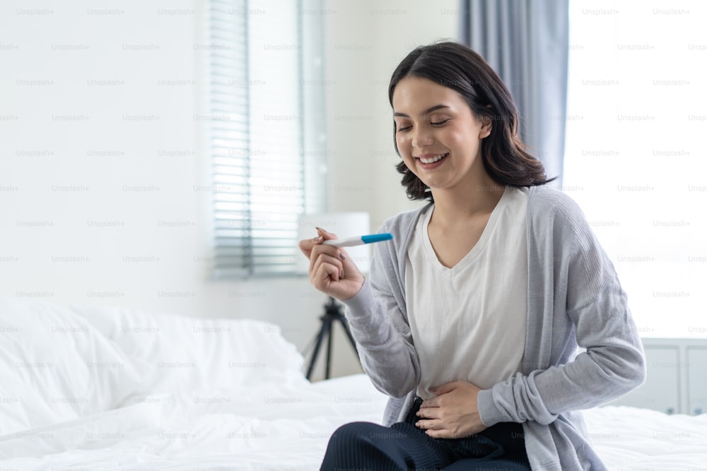 Asian young woman feeling happy after look at positive pregnancy test. Beautiful loving female feel happy and excited for good news to be future parent on bed in bedroom. Family-Expecting baby concept