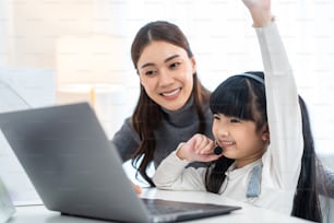 Asian little young girl kid learning online class at home with mother. Preschool child use laptop computer do homework, homeschool from school teacher by digital remote internet with support from mom.