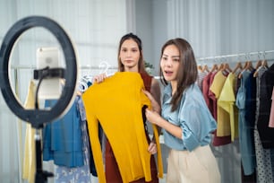 Asian woman friends couple talk to camera to sell the clothes at home. Young attractive business people female live streaming and showing clothing to customer in online retail to sales at office store