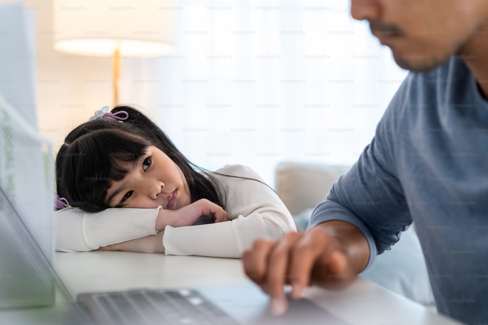 Asian young kid daughter feeling sad and need to play with busy father. Attractive parent daddy use laptop computer working from home during new normal and have no time for little child girl in house.