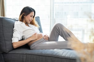 自宅のソファに座って両手でおなかを抱えている若いアジアの妊娠中の女性。妊娠中の女の子はお腹を見ます。お腹の中の赤ちゃんの触感。将来、生まれたばかりの赤ちゃんを出産することを考えて