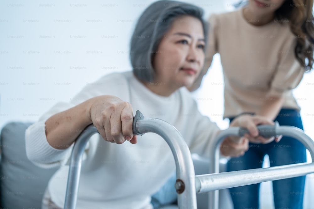Une jeune fille asiatique soutient une vieille femme handicapée qui marche avec un déambulateur à la maison. Belle fille aide et prend soin d’une personne âgée âgée mature handicapée maman patient faisant de la physiothérapie dans le salon dans la maison