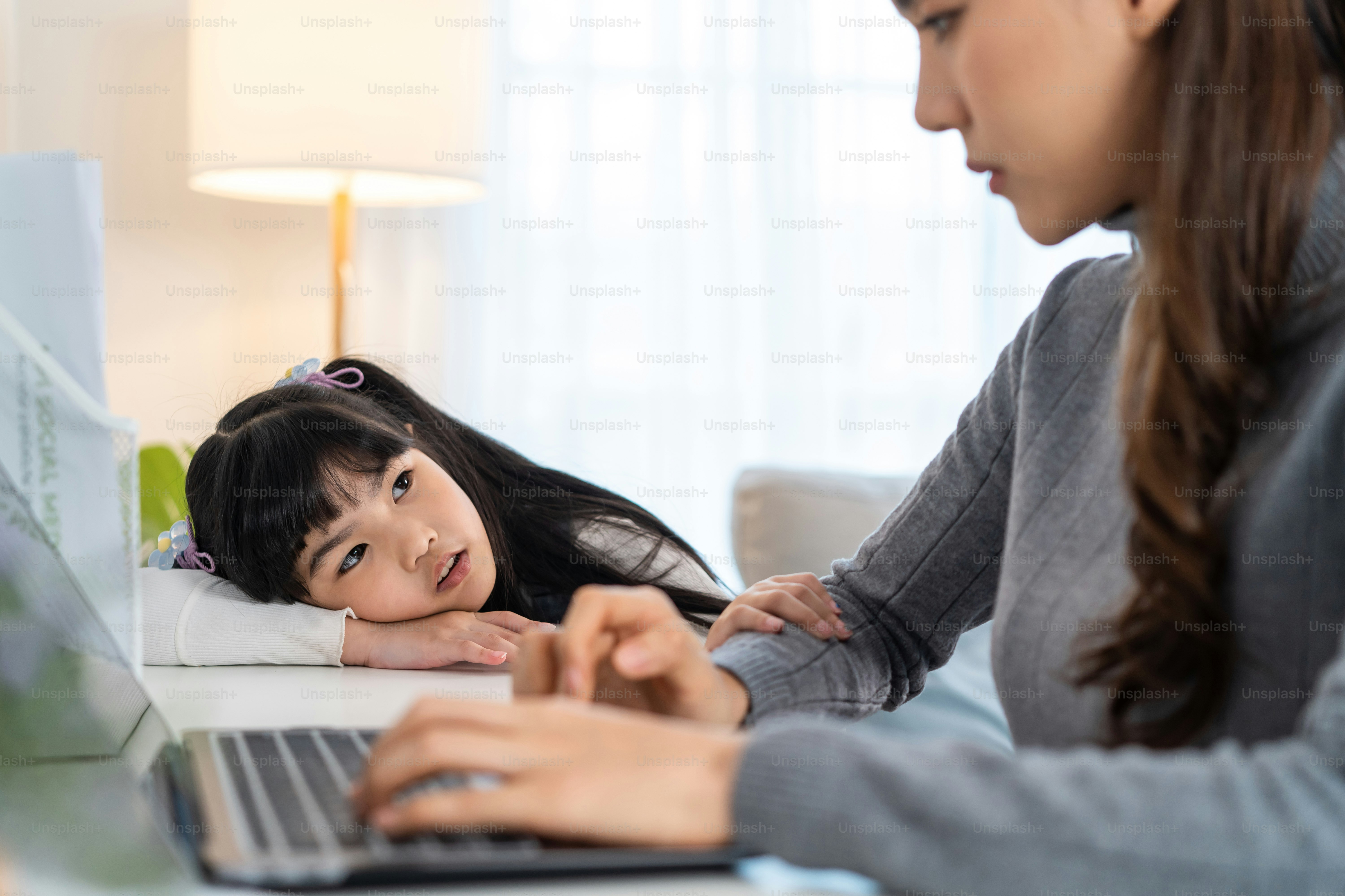 Asian young kid daughter feeling sad and need to play with busy mother. Attractive beautiful mom use laptop computer working from home during new normal and have no time for little child girl in house