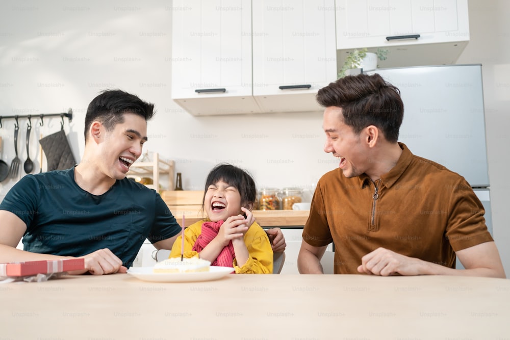 Asian attractive LGBTQ gay family celebrate birthday girl kid in house. Handsome male couple take care and surprise little adorable girl child with cake and present gift on her special day at home.