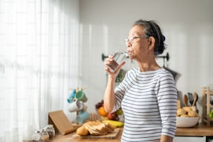 Asian senior mature woman drinking a glass of milk in kitchen at home. Attractive elderly older grandmother holding and sipping a cup of milk after waking up in the morning for health care in house.