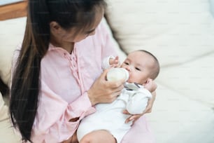 Asiatische junge Mutter, die süßes Neugeborenes aus der Milchflasche im Schlafzimmer hält und füttert. Mädchen kümmert sich um süßen Jungen mit warmem Lächeln. Glücklicher Junge bleibt zusammen mit seiner Mutter in einem schönen Zuhause. Neugeborenen-Baby-Familienkonzept.