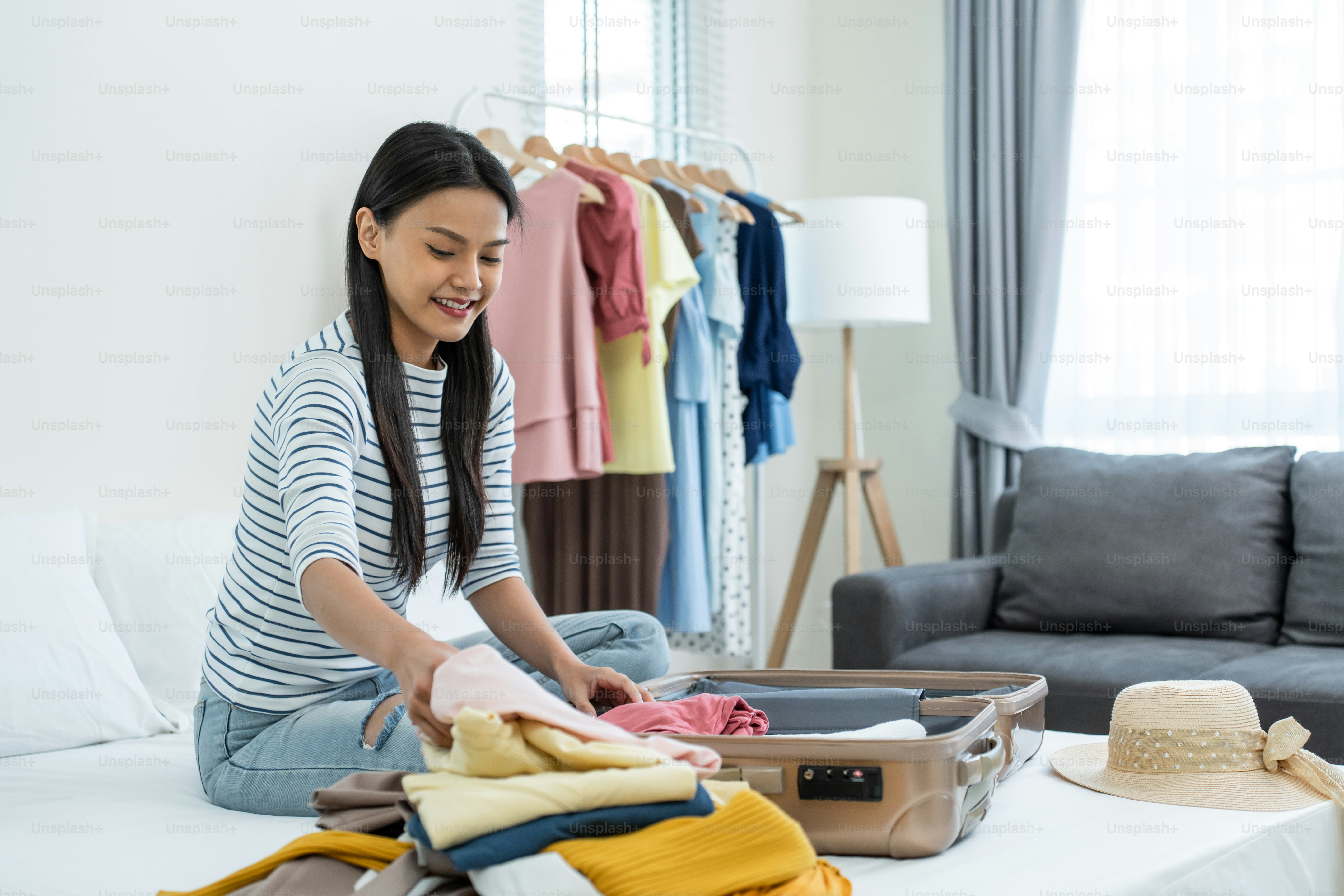 Asian young beautiful woman preparing clothes and packing a suitcase. Attractive female tourist traveler feel happy and relax while preparing luggage on bed, ready to travel on holiday vacation trip.