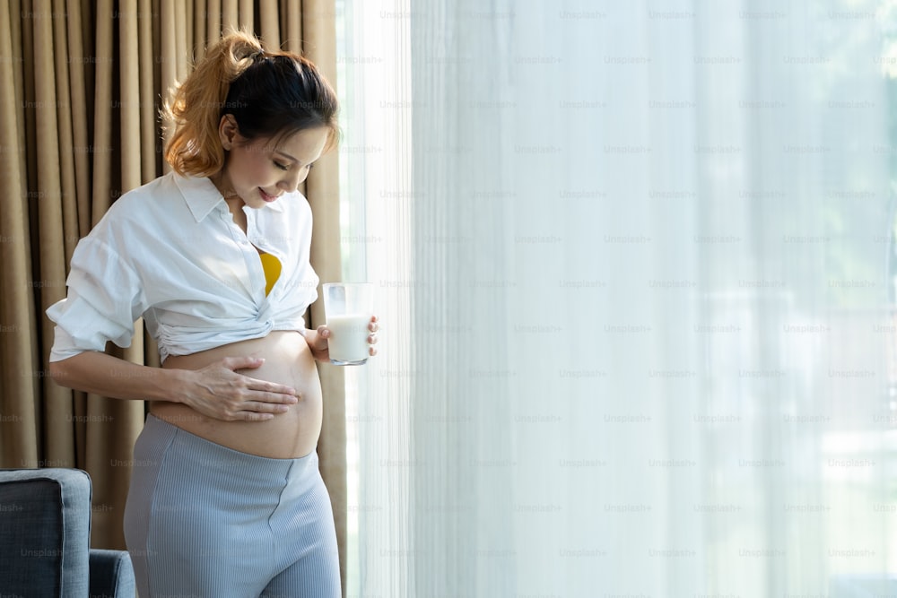 Asian beautiful pregnancy woman drink a glass of milk for health care. Attractive pregnant girl stand indoors in living room and touching and stroking baby in the womb with happiness and love in house