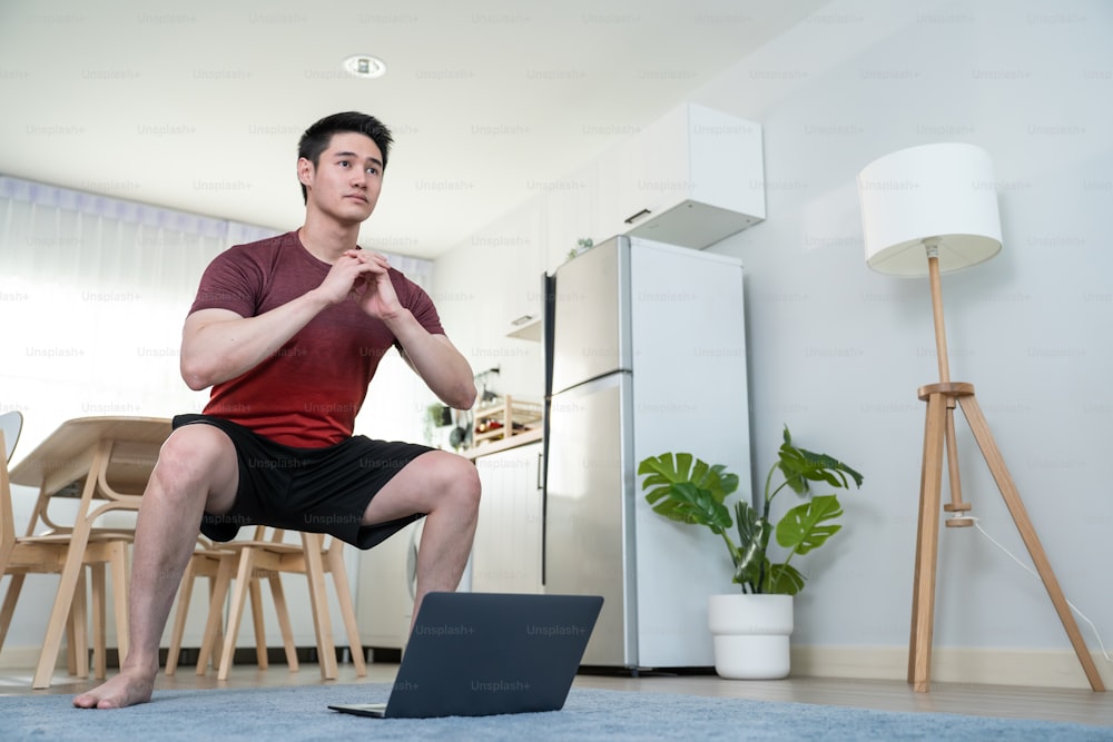 Asian attractive young man doing exercise using laptop in living room. Handsome male in sport cloth follow exercise or yoga instruction video online trainer workout at home. Healthy lifestyle concept.